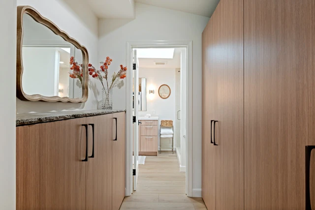 Dressing vestibule with full-height Pronorm wardrobe and a low fluted-front credenza below a wavy hand-carved mirror, looking through to the primary bath